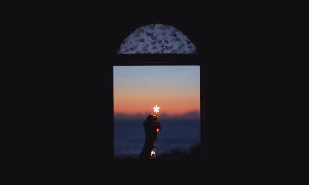 Hand holding a small glowing star against a sunset sky, framed by a dark background, symbolizing hope