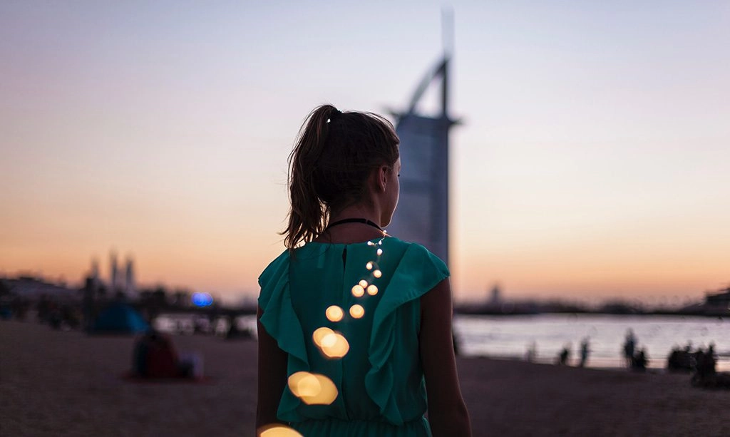 A person seen from behind stands by a waterfront at dusk, looking toward a softly lit city skyline as warm bokeh lights trail across the foreground.